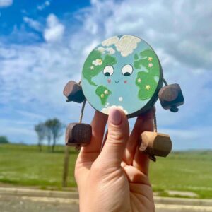 Handmade wooden toy shaped like the Earth, hand painted with continents, clouds and small flowers, with a smiling face. Made from natural oak, beech or walnut with rope limbs and wooden feet, designed for open-ended play and nature-inspired storytelling.