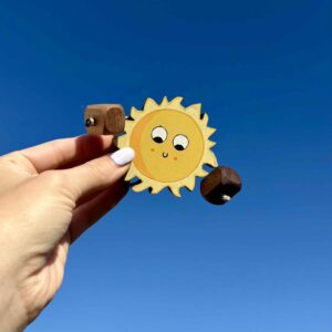 Handmade wooden sun toy (Sally Sun) held in a hand against a clear blue sky, made from natural oak, beech or walnut with hand painted sun details and a smiling face. The toy has rope limbs with wooden hands and is designed for open-ended play and imaginative storytelling.