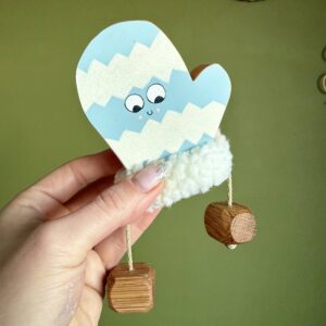 Hand holding a wooden winter mitten decoration with a blue and cream zigzag design, smiling face, fluffy white cuff and dangling wooden legs