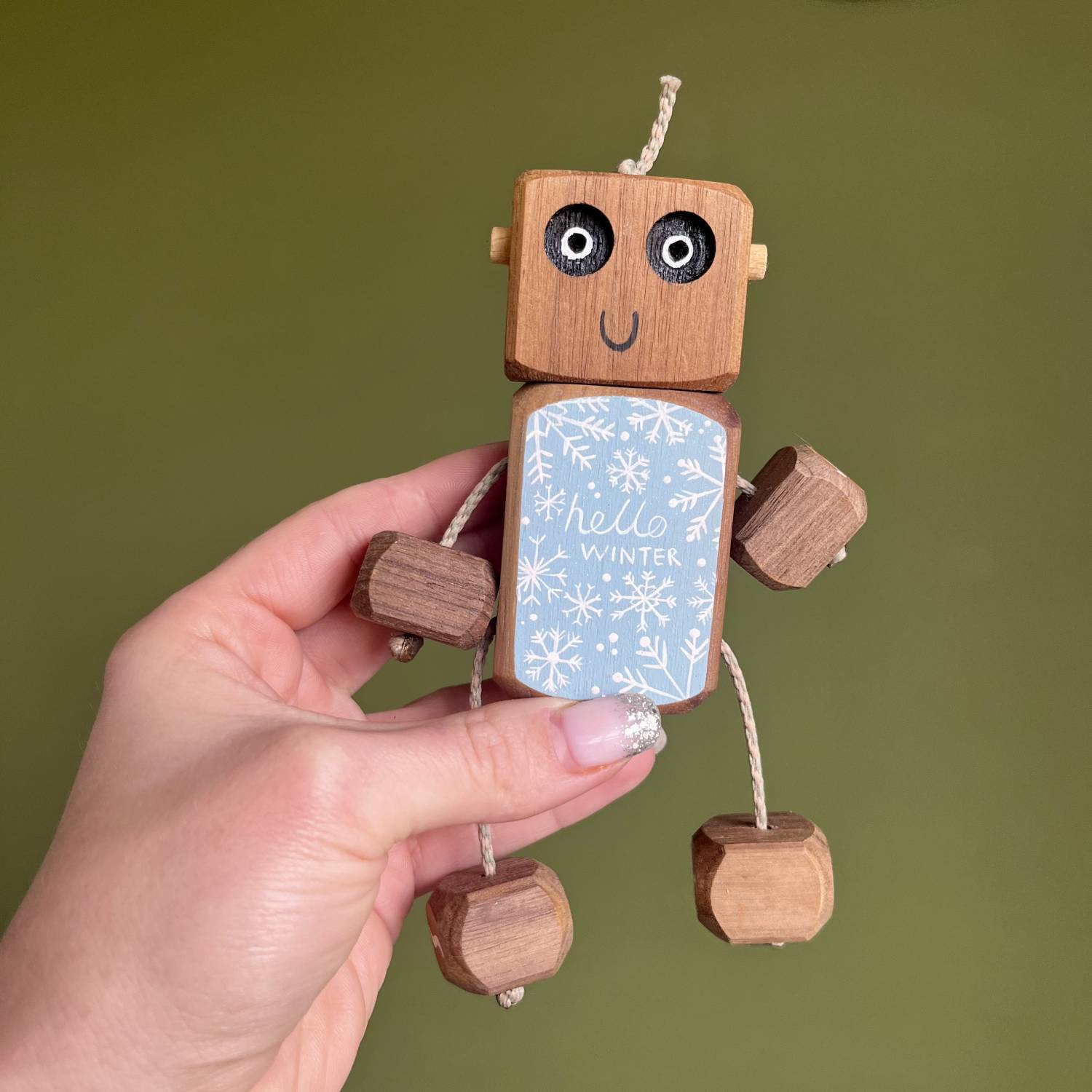 Hand holding a small handcrafted wooden robot toy with a blue “hello winter” snowflake design on its body, made from walnut wood with string limbs.