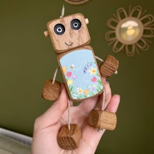 Handmade wooden robot toy (Ned) held in a hand, made from natural oak or walnut and hand painted with colourful spring flowers and a small bee on a soft blue background. The toy has moveable rope arms and legs and is designed for open-ended play and keepsake gifting.