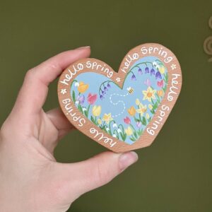 Handmade wooden heart held in a hand, hand painted with colourful spring flowers including tulips, daffodils and bluebells, with a small bee detail on a soft blue background. The wooden heart reads “Hello Spring” around the edge and is crafted from natural wood, designed as a unique spring decoration or keepsake gift.