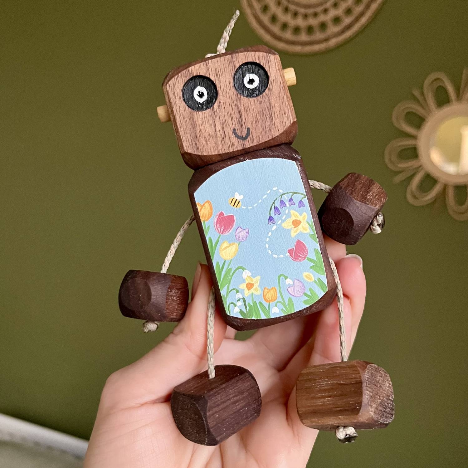 Handmade wooden robot toy (Ned) held in a hand, made from natural oak or walnut and hand painted with colourful spring flowers and a small bee on a soft blue background. The toy has moveable rope arms and legs and is designed for open-ended play and keepsake gifting.