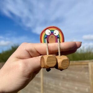 Hand holding a small handmade wooden rainbow fidget toy with a smiling face and dangling wooden feet, designed for sensory play and open-ended play, photographed against a bright blue sky