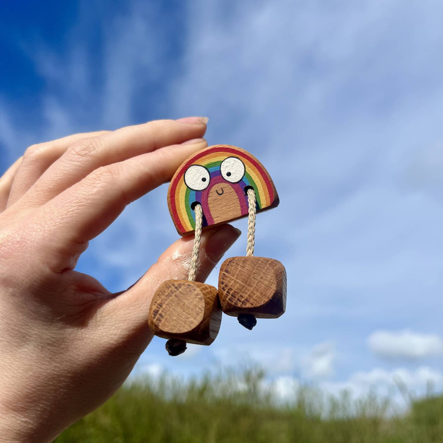Hand holding up a small handmade wooden rainbow fidget toy with a smiling face and dangling wooden feet, designed for sensory play and open-ended play, photographed against a bright blue sky
