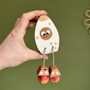 Handmade wooden rocket toy (Rocket Ned) held in a hand, made from natural oak, beech or walnut with hand painted details and a small smiling face. The toy has rope legs with wooden feet and is designed for open-ended play, inspired by space and adventure.